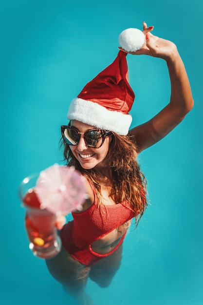 Joven mujer hermosa en la piscina con sombrero de Santa Claus, celebra Año Nuevo y Navidad en un país cálido con una copa de cóctel.