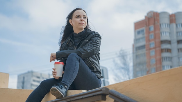 Una joven mujer hermosa con ropa informal está sentada en una valla de hormigón y tomando un café al fondo de la ciudad