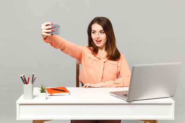 Joven mujer hermosa con ropa pastel haciendo selfie en el teléfono móvil, trabaja en el escritorio con una computadora portátil aislada en un fondo gris. Concepto de estilo de vida de carrera empresarial de logro. Simulacros de espacio de copia.