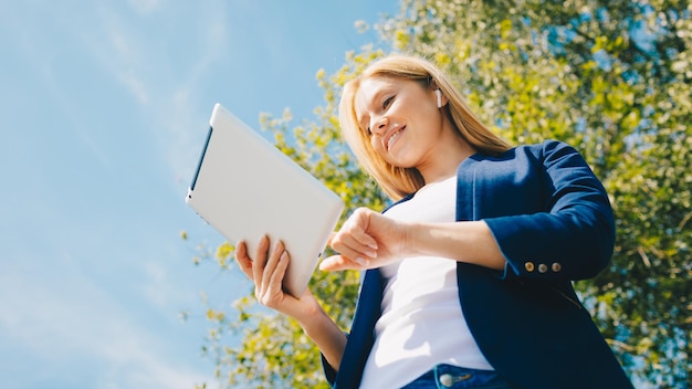 Joven mujer hermosa sonriente en el parque con un dispositivo de tableta y auriculares inalámbricos disfrutando del trabajo remoto del concepto de energía verde