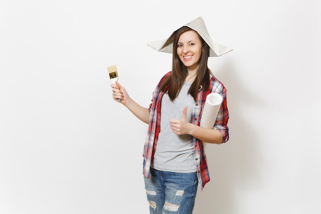 Joven mujer hermosa sonriente con sombrero de periódico sosteniendo rollo de papel tapiz y pincel y mostrando el pulgar hacia arriba aislado sobre fondo blanco. Instrumentos para la renovación de la habitación del apartamento. Reparar el concepto de hogar.