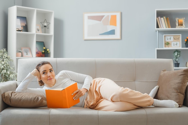 Joven mujer hermosa vestida de casa relajándose en un sofá en el interior de su casa leyendo un libro feliz y positiva mirando a un lado con una expresión soñadora pasando el fin de semana en casa