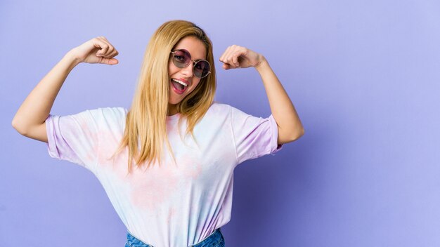 Joven mujer hipie con gafas aisladas en púrpura mostrando gesto de fuerza con los brazos, símbolo del poder femenino