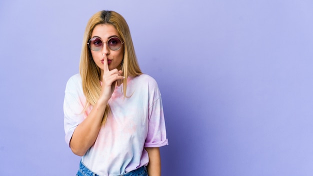 Joven mujer hippie con gafas en la pared púrpura guardando un secreto