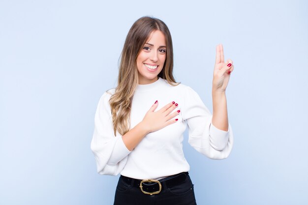 Joven mujer hispana bonita que parece feliz, confiada y confiable, sonriendo y mostrando el signo de la victoria, con una actitud positiva contra la pared azul