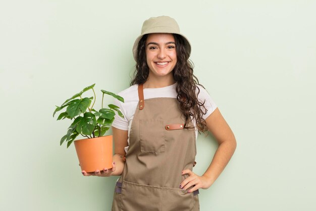 Joven mujer hispana bonita sonriendo felizmente con una mano en la cadera y un concepto de agricultor o jardinero confiado