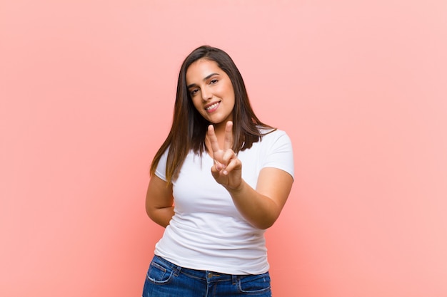 Joven mujer hispana bonita sonriendo y mirando feliz, despreocupada y positiva, gesticulando victoria o paz con una mano contra la pared rosa