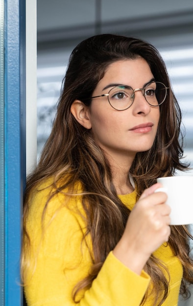 Joven mujer hispana en una camisa amarilla bebiendo café y mirando por la ventana