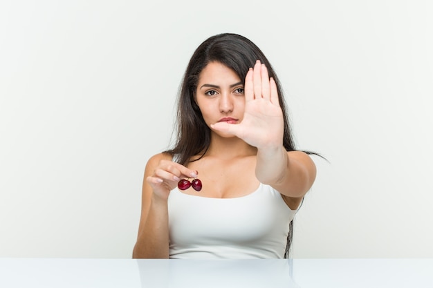 Joven mujer hispana con cerezas de pie con la mano extendida que muestra la señal de stop, impidiéndole.