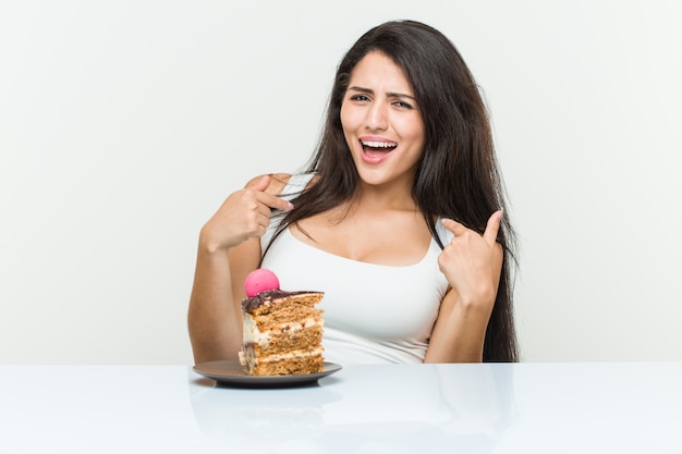 La joven mujer hispana comiendo un pastel sorprendida señalando algo a sí mismo, sonriendo ampliamente.