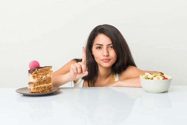 Joven mujer hispana elegir entre pastel o fruta mostrando número uno con el dedo.