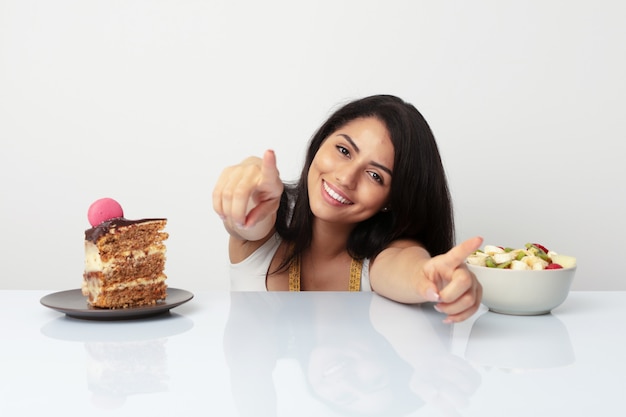 Joven mujer hispana elegir entre pastel o fruta sonrisas alegres apuntando al frente.