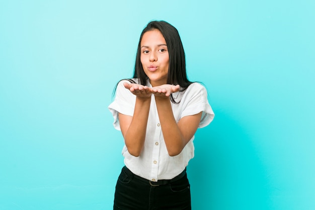 Joven mujer hispana fresca contra una pared azul doblando los labios y sosteniendo las palmas para enviar beso al aire.