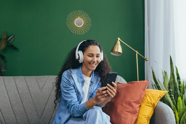 Joven mujer hispana sonriente en casa con auriculares y teléfono escuchando música en línea usando