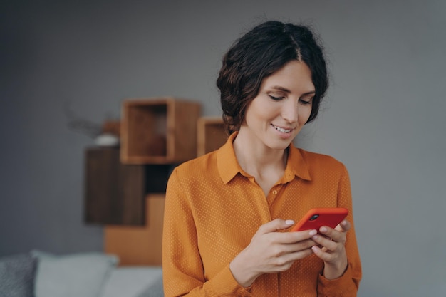 Joven mujer hispana sonriente sosteniendo un teléfono inteligente mirando la pantalla con una sonrisa y enviando mensajes de texto