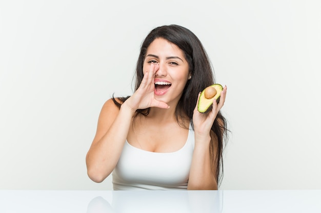 Joven mujer hispana sosteniendo un aguacate gritando emocionado al frente.