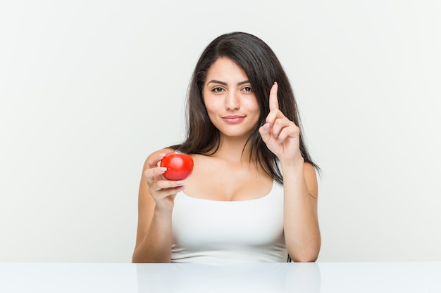 Joven mujer hispana sosteniendo un tomate mostrando el número uno con el dedo.