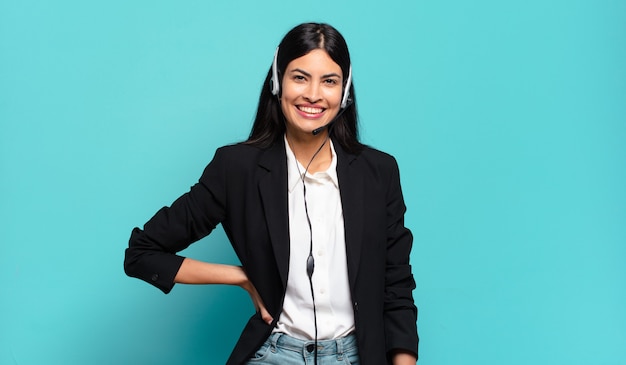 Joven mujer hispana de telemercadeo sonriendo felizmente con una mano en la cadera y una actitud segura, positiva, orgullosa y amistosa