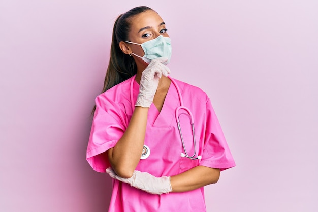 Joven mujer hispana con uniforme de médico y máscara médica sonriendo mirando con confianza a la cámara con los brazos cruzados y la mano en la barbilla pensando positivamente