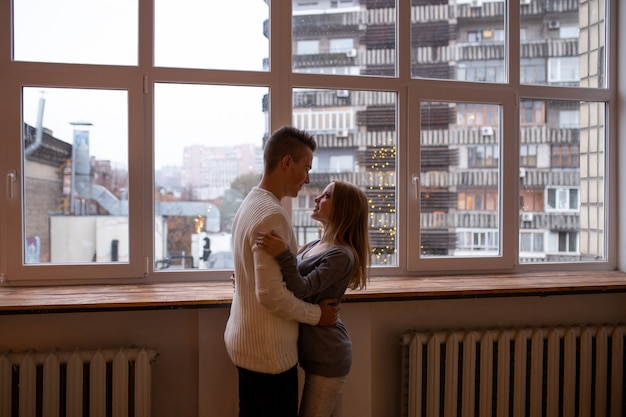 Joven mujer y hombre enamorado se abrazan de pie cerca de la ventana en casa