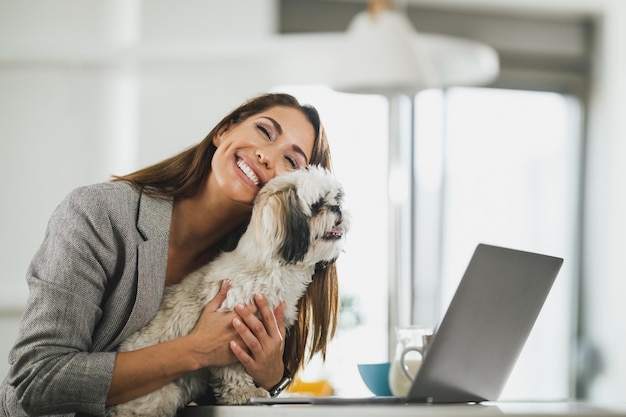 Joven mujer independiente sentada con su perro mascota y usando su laptop durante el trabajo desde casa.