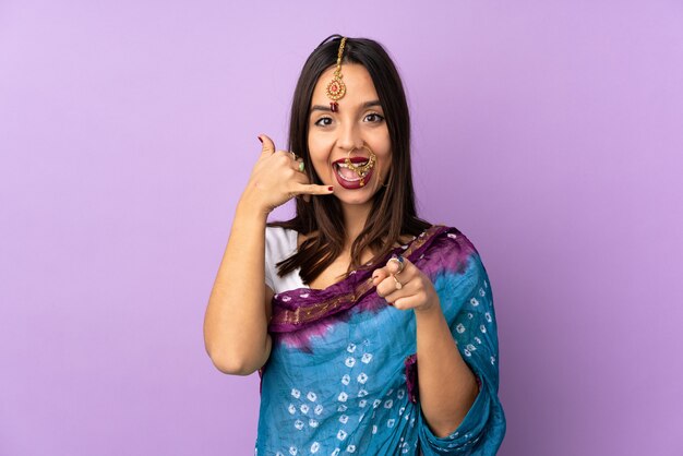 Joven mujer india aislada en púrpura haciendo gesto de teléfono y apuntando hacia delante