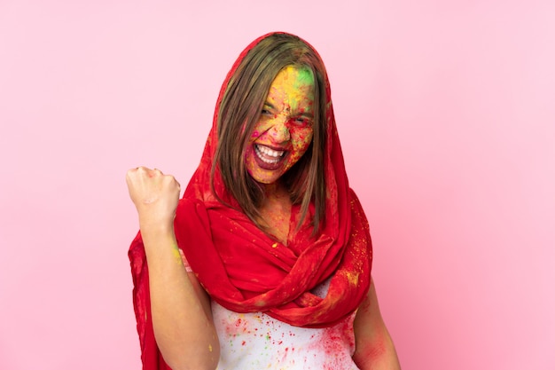 Joven mujer india con coloridos polvos de holi en la cara en la pared rosa celebrando una victoria