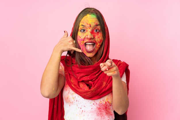 Joven mujer india con coloridos polvos de holi en su rostro aislado en la pared rosa haciendo gesto de teléfono y apuntando hacia el frente