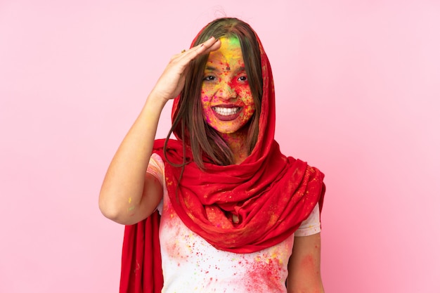 Joven mujer india con coloridos polvos de holi en su rostro aislado en la pared rosa saludando con la mano con expresión feliz