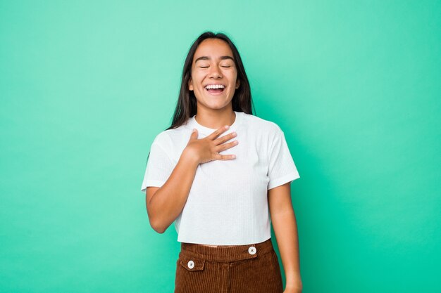 Joven mujer india de raza mixta aislada se ríe a carcajadas manteniendo la mano en el pecho.