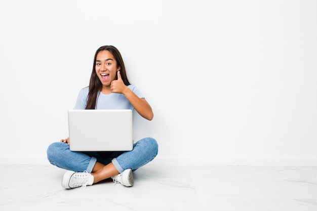 Joven mujer india de raza mixta sentada trabajando en la computadora portátil sonriendo y levantando el pulgar hacia arriba