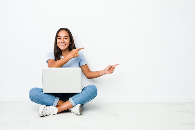 Joven mujer india de raza mixta sentado trabajando en la computadora portátil emocionado apuntando con los dedos índices.