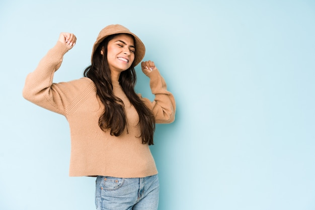 Joven mujer india con un sombrero en la pared azul levantando el puño después de una victoria