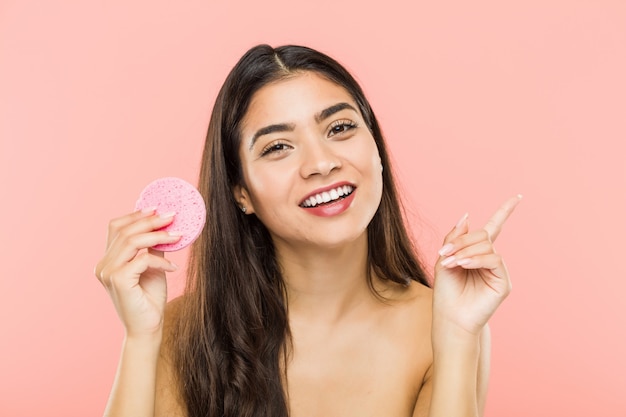 Joven mujer India sosteniendo un disco de cuidado de la piel facial sonriendo alegremente apuntando con el dedo lejos.