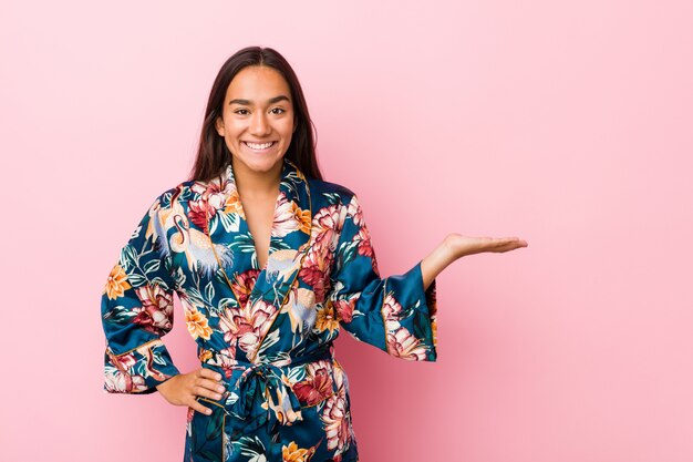 Joven mujer India vistiendo un pijama kimono mostrando un espacio de copia en una palma y sosteniendo otra mano en la cintura.