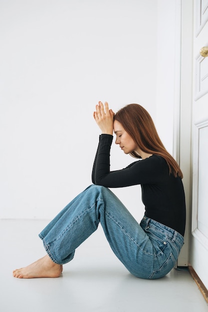 Joven mujer infeliz adolescente con cabello largo en jeans sentada en el piso en posición cerrada junto a la puerta en casa emoción negativa