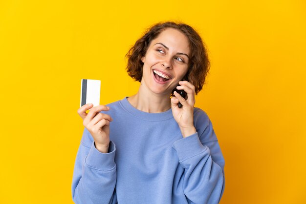 Joven mujer inglesa aislada en el espacio amarillo manteniendo una conversación con el teléfono móvil y sosteniendo una tarjeta de crédito