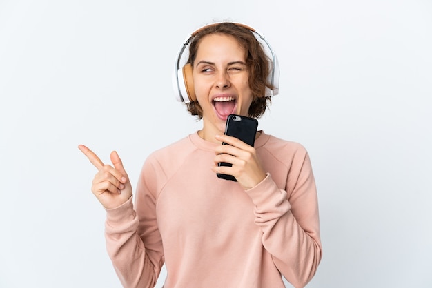 Joven mujer inglesa aislada en la pared blanca escuchando música con un móvil y cantando