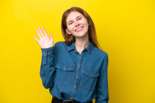 Joven mujer inglesa aislada sobre fondo amarillo saludando con la mano con expresión feliz