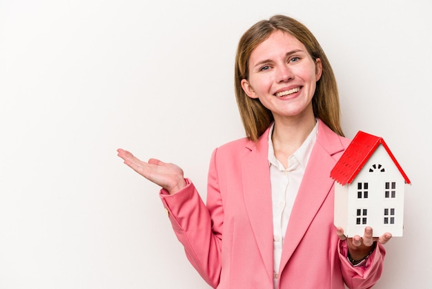 Joven mujer inglesa de negocios sosteniendo un juguete de la casa aislado de fondo blanco mostrando un espacio de copia en una palma y sosteniendo otra mano en la cintura