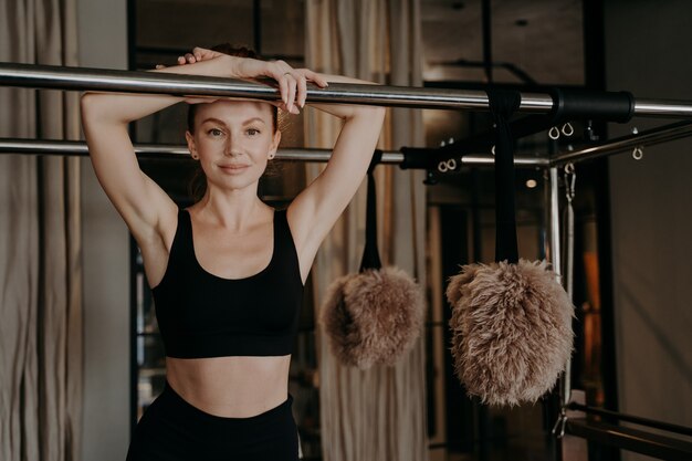 Joven mujer de jengibre feliz en ropa deportiva negra tomando un descanso en la máquina de pilates entre entrenamientos en el gimnasio, mirando con alegría sonriendo a la cámara, estilo de vida saludable y concepto de deporte en el interior