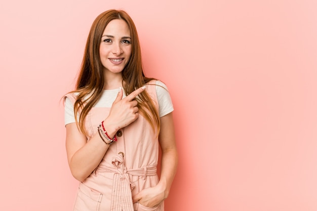 Joven mujer de jengibre con pecas sonriendo y apuntando a un lado, mostrando algo en el espacio en blanco.