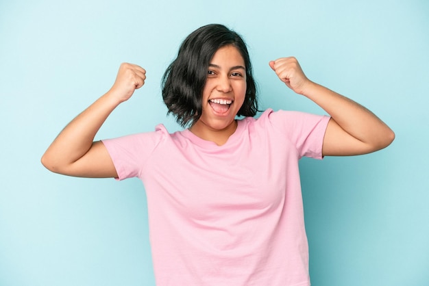 Joven mujer latina aislada sobre fondo azul mostrando gesto de fuerza con los brazos, símbolo del poder femenino