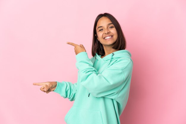 Joven mujer latina aislada sobre fondo rosa apuntando con el dedo hacia un lado y presentando un producto