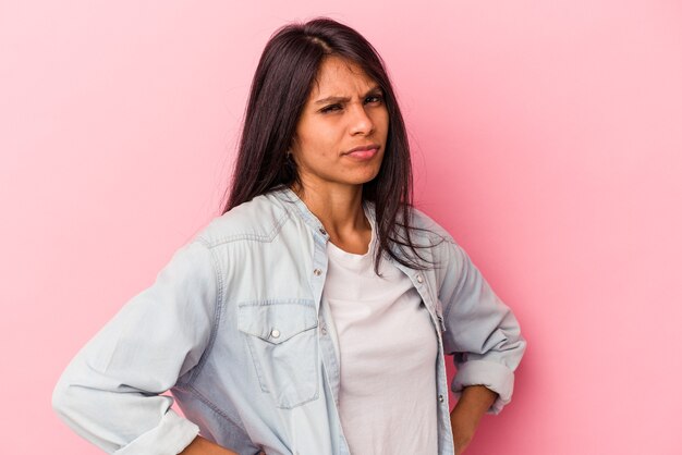 Joven mujer latina aislada sobre fondo rosa con el ceño fruncido en disgusto, mantiene los brazos cruzados.