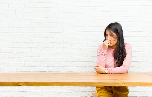 Joven mujer latina bonita que se siente disgustada, tapándose la nariz para evitar oler un hedor desagradable y desagradable sentado frente a una mesa