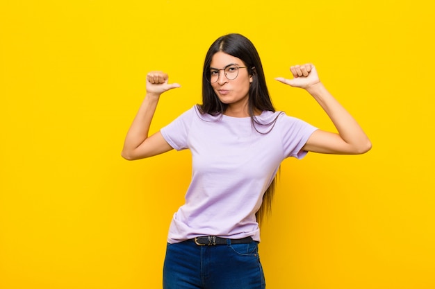 Joven mujer latina bonita que se siente orgullosa, arrogante y segura de sí misma, luciendo satisfecha y exitosa, apuntando a sí misma contra la pared plana