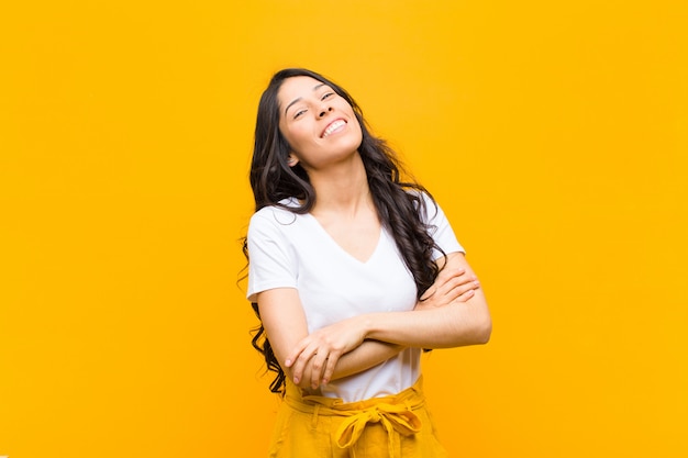 Joven mujer latina bonita riendo alegremente con los brazos cruzados, con una pose relajada, positiva y satisfecha contra la pared naranja