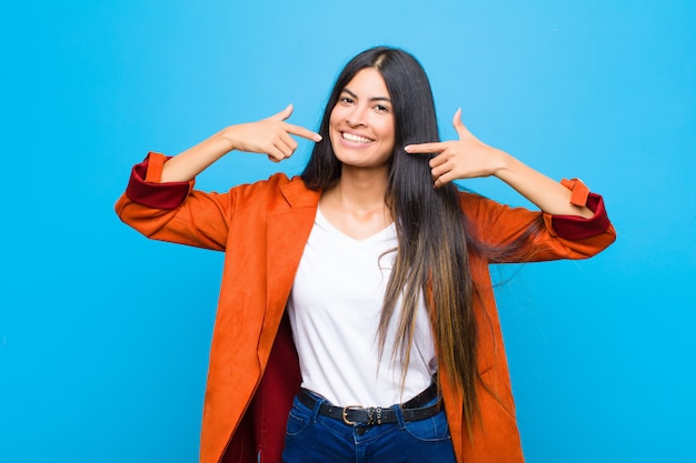 Joven mujer latina bonita sonriendo con confianza apuntando a su propia sonrisa amplia, actitud positiva, relajada y satisfecha contra la pared plana