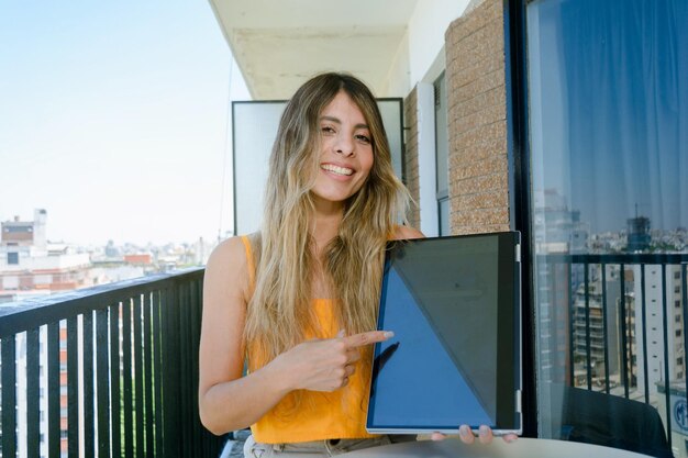 Joven mujer latina colombiana riéndose en el balcón de su apartamento mostrando la pantalla de su computadora portátil